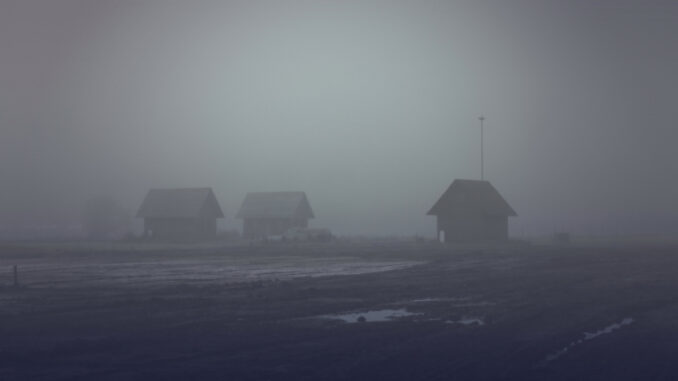 The wooden houses on a dark background. A fog on the edge of the forest and fields. The landscape of horror
