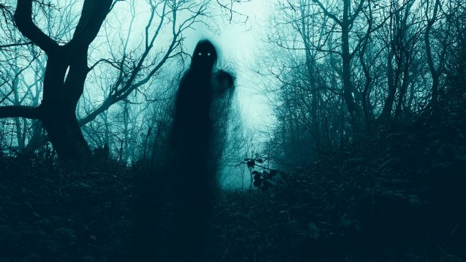 A horror concept of a blurred scary hooded figure with glowing eyes on a path through a moody misty winter woodland